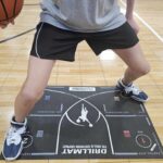 A certified I'm Possible trainer guides a person practicing basketball footwork on a skill training mat, holding a basketball on the gym floor.